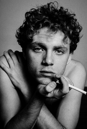 Black and white picture of a white man with curly hair staring into the camera holding a cigarette in his hand. This illustrates how grieving is painful and hard. Sacred Circle Holistic Healing offers online therapy in Kansas for grief and loss.