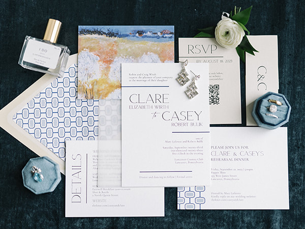 white invitation  and details cardwith blue foil pressed accent stripes, Details card with painting of a country scene,  Reply card on tan paper, Second invitation with retro patternl white envelope with repeated retro pattern liner
