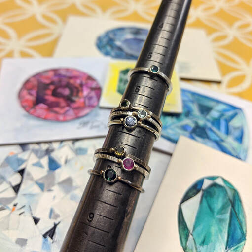 assorted rings on a ring mandrel with paintings of stones in the background