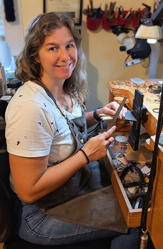 A woman sits at a jewelry bench holding a file and smiling at the camera