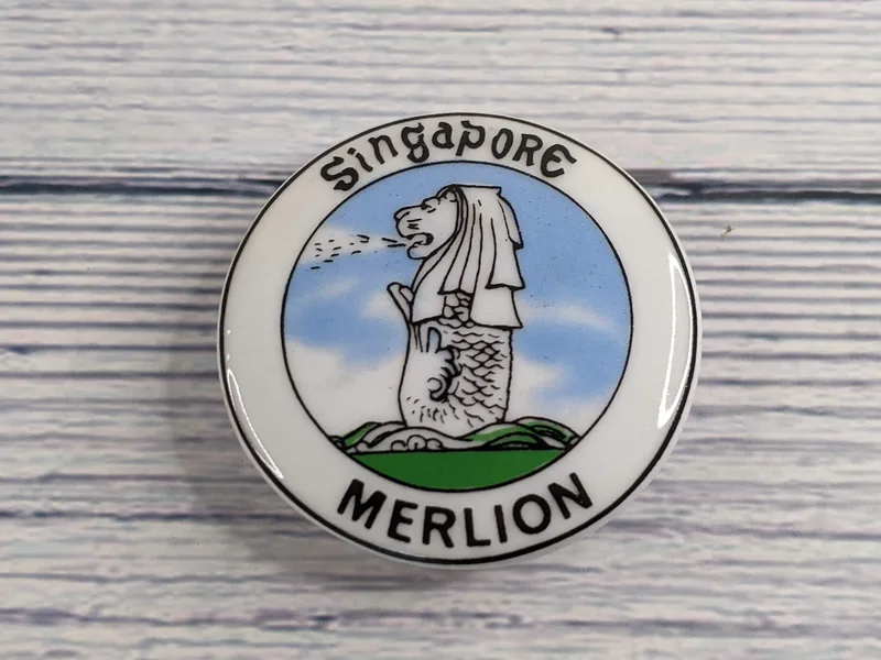 A porcelain magnet that is dome shaped with an image of the Singapore Merlion statue.