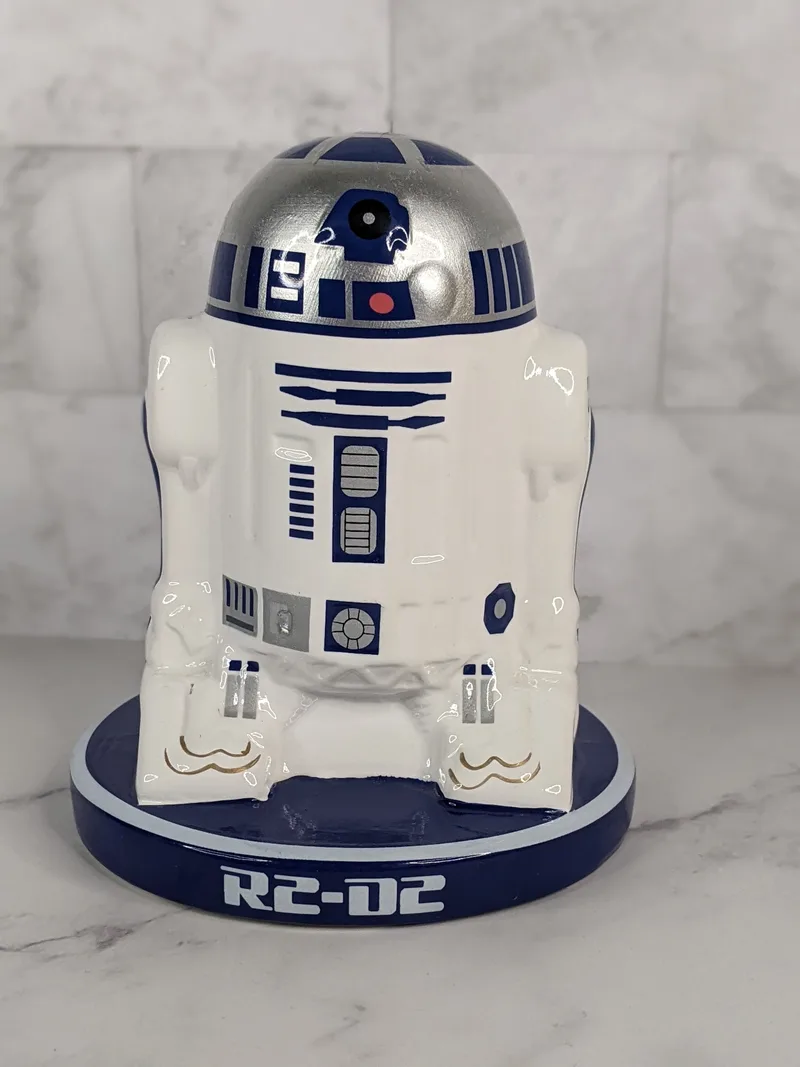 A Star Wars themed coin bank in the likeness of the R2-D2 character, the white, blue, and silver robot.
