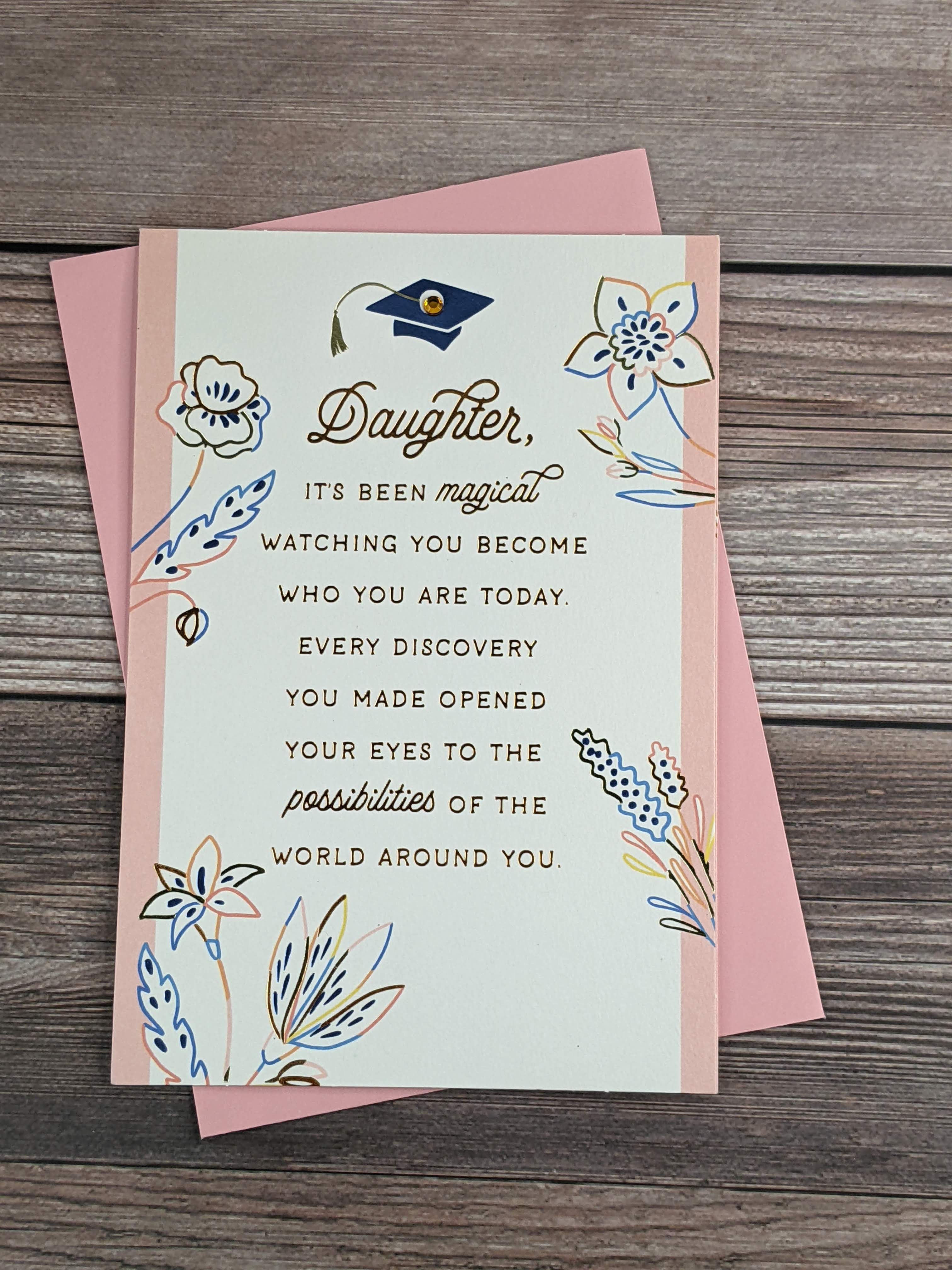 A graduation card for a daughter, featuring a blue grad cap with a yellow gem, a flower design with pink accents, and a pink envelope.