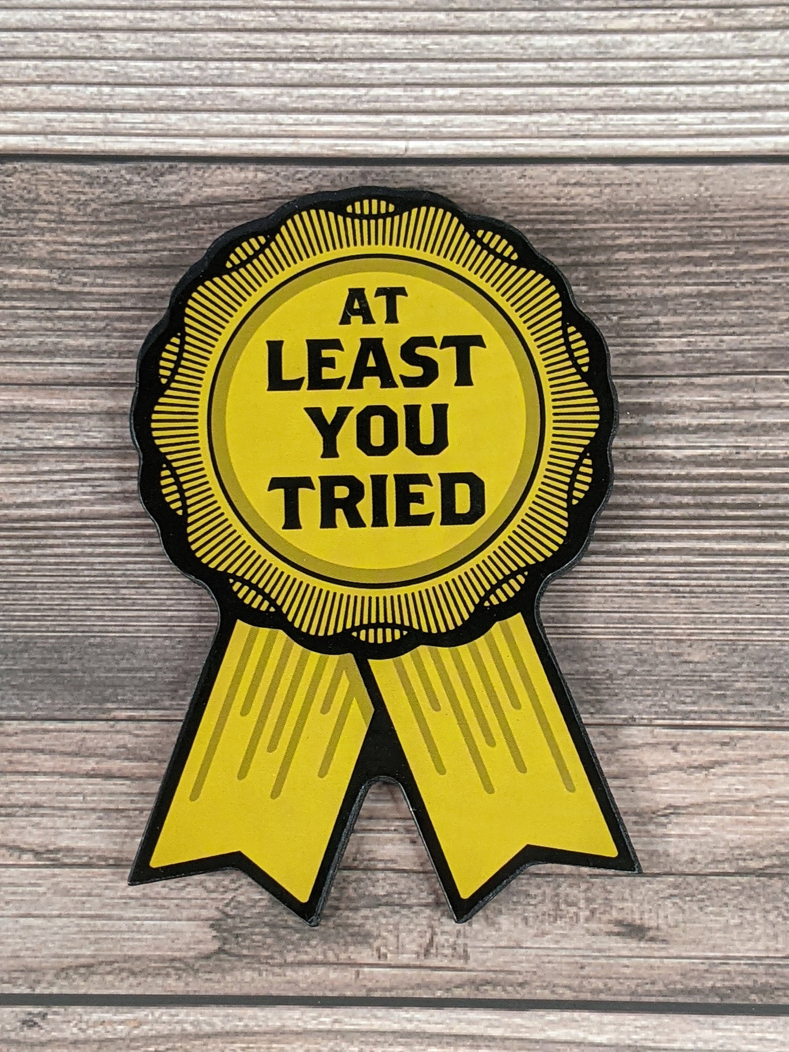A yellow magnet shaped like a ribbon award with black lettering of the words, "at least you tried."