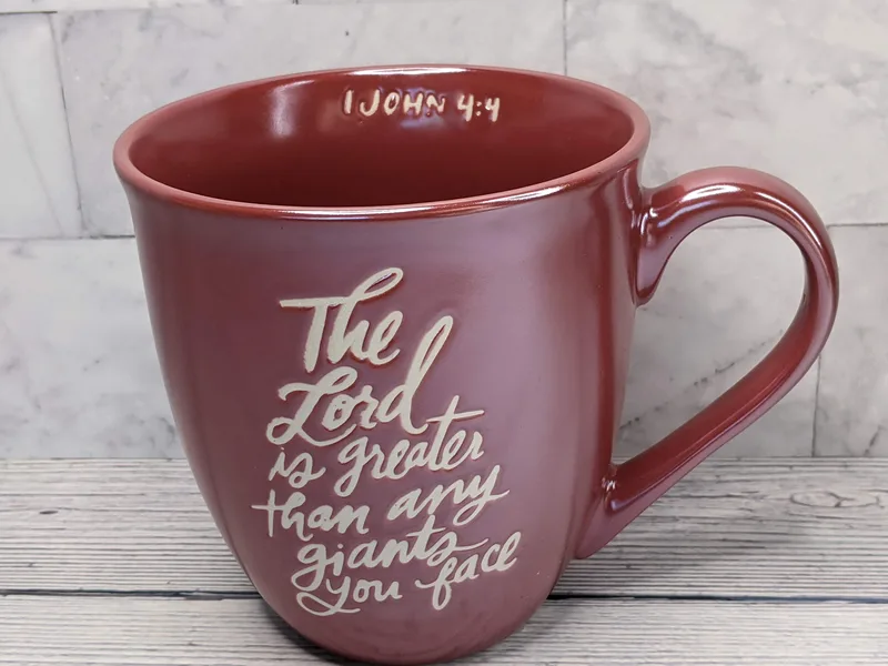 A red mug with tan lettering of the words "The Lord is greater than any giants you face. -1 John 4:4"