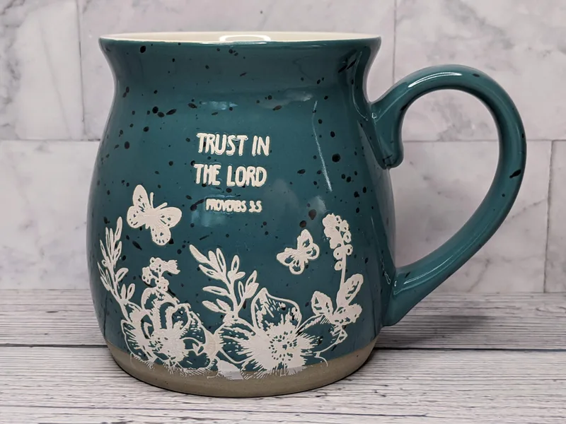 A large teal coffee mug with a Bible verse, Trust in the Lord, and a stamped image of flowers and butterflies.