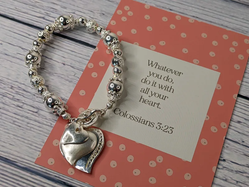 A Bible verse card with a beaded bracelet that has heart charms and silver metal beads. Colossians 3:23