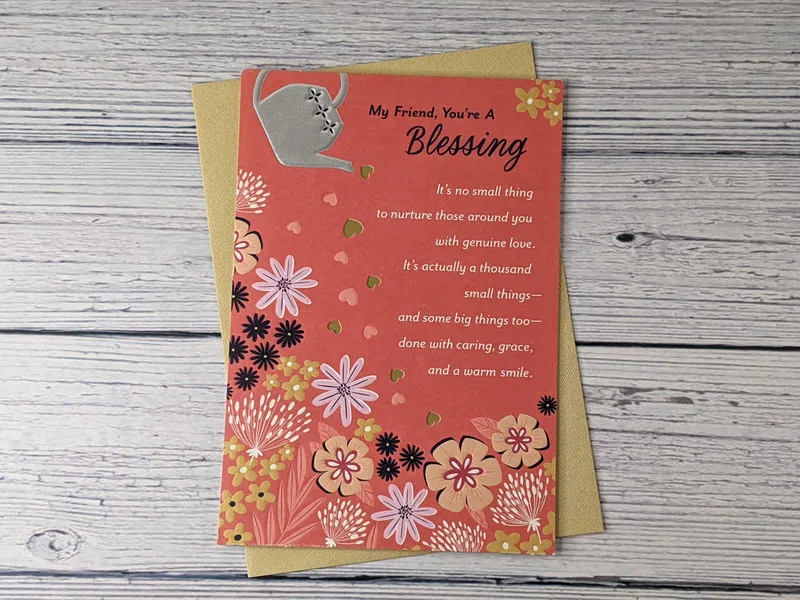 A coral cover of a friend birthday greeting card with an image of a silver watering can pouring hearts onto flowers.