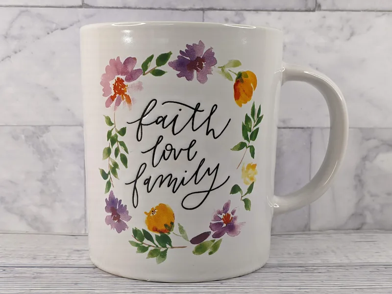 A ceramic white mug with a lavender interior. There is a circular flower design (purple and orange) on the front with the words "faith, love, family" in a cursive font.