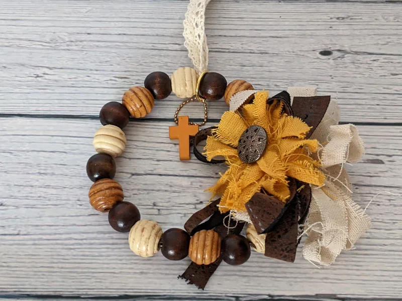 A wooden beaded wreath ornament with a cross charm and shabby fabric bow and golden sunflower.