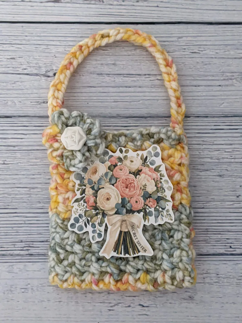 A yellow and green crocheted gift bag with a handle and a secret sister gift tag.