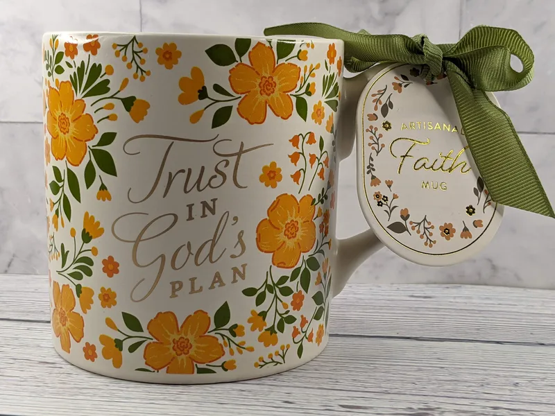 A tan mug with an orange floral design and gold lettering with the words Trust in God's plan.
