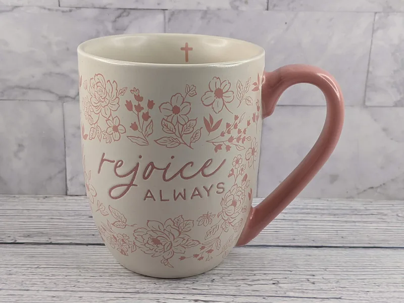 A tan mug with a pink design of flowers and the words "rejoice always."
