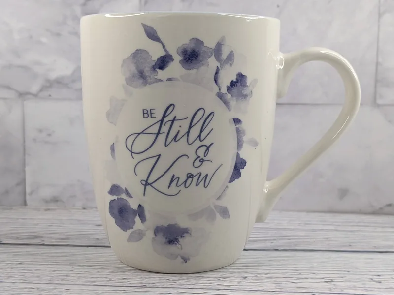 A ceramic white mug with blue flowers and the words "be still & know."