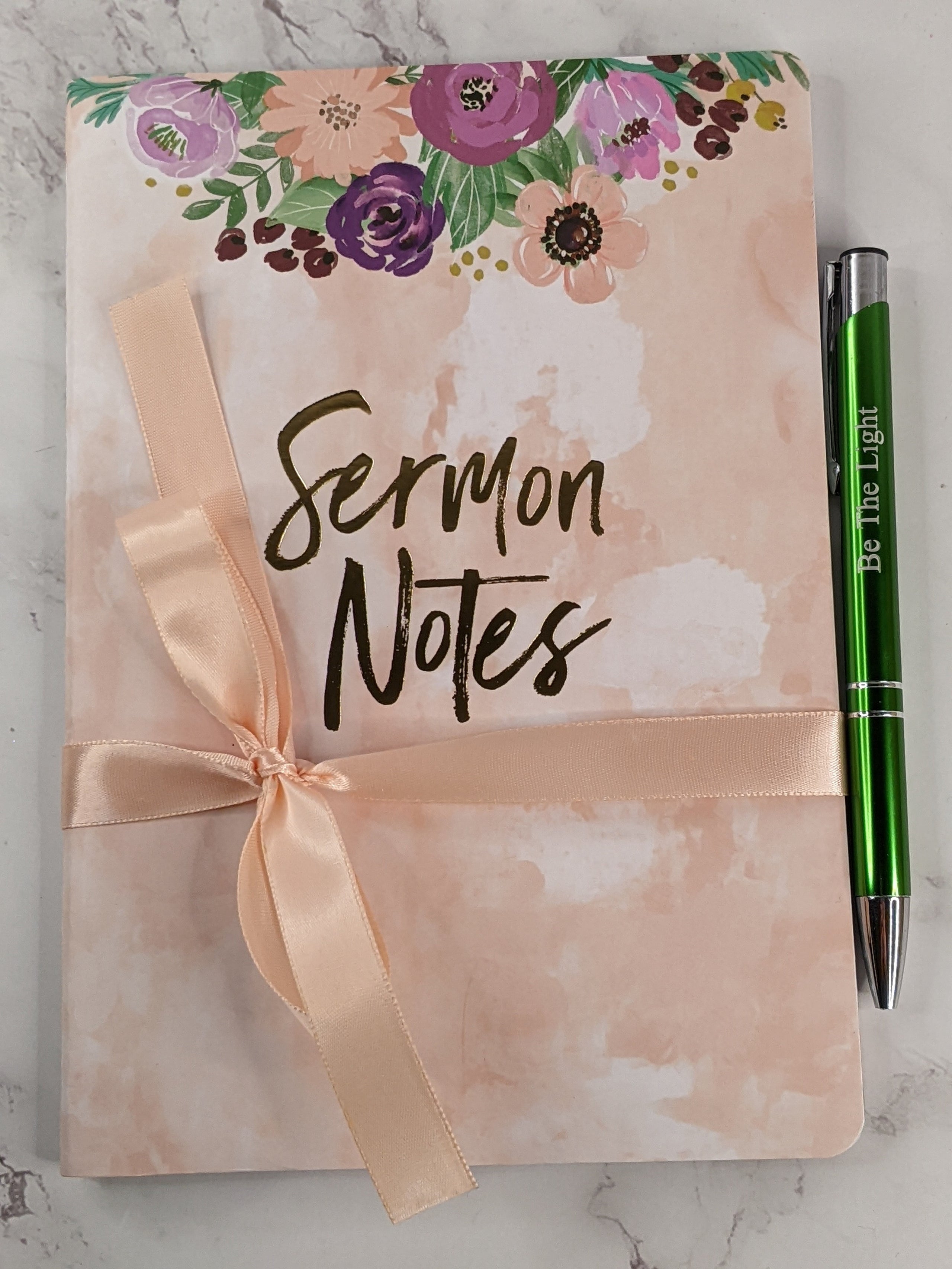 A flower designed cover on a religious journal with lined pages for writing sermon notes, and a green "Be the Light" pen.