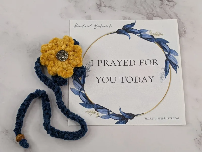 A crocheted plush yarn yellow flower bookmark with a blue strand and tassel, along with a "I prayed for you today" religious message card.