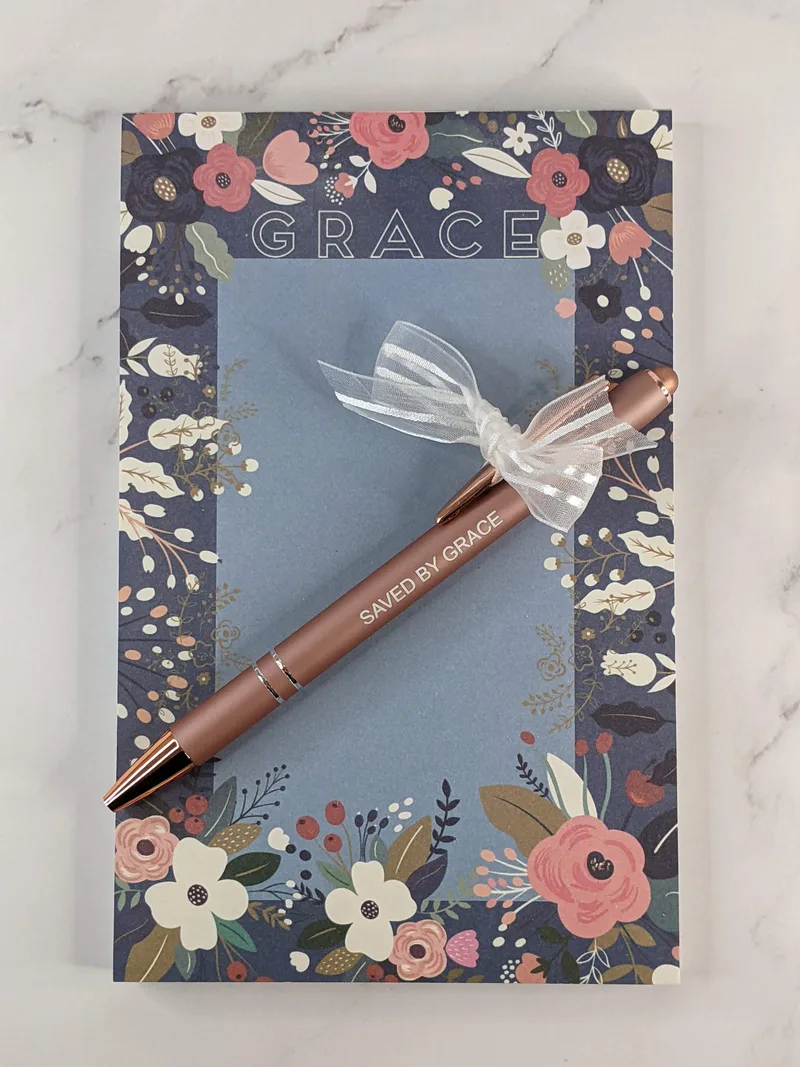 A medium-sized 7" x 4.5" blue GRACE Notepad with a floral design and a mauve Saved By Grace Pen.