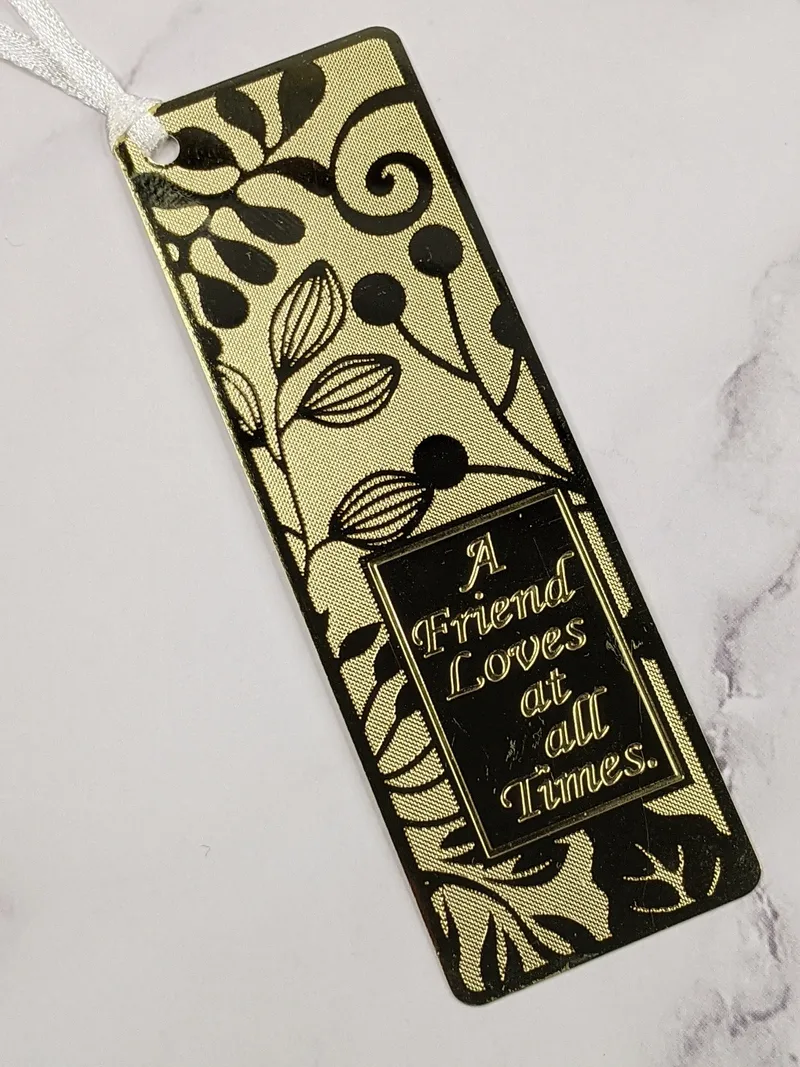 A gold metal bookmark with an embossed leaf design and an inscription, "A friend loves at all times."