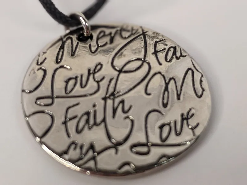 A black-cord necklace with a silver metal pendant featuring the words Love, Faith and Mercy.