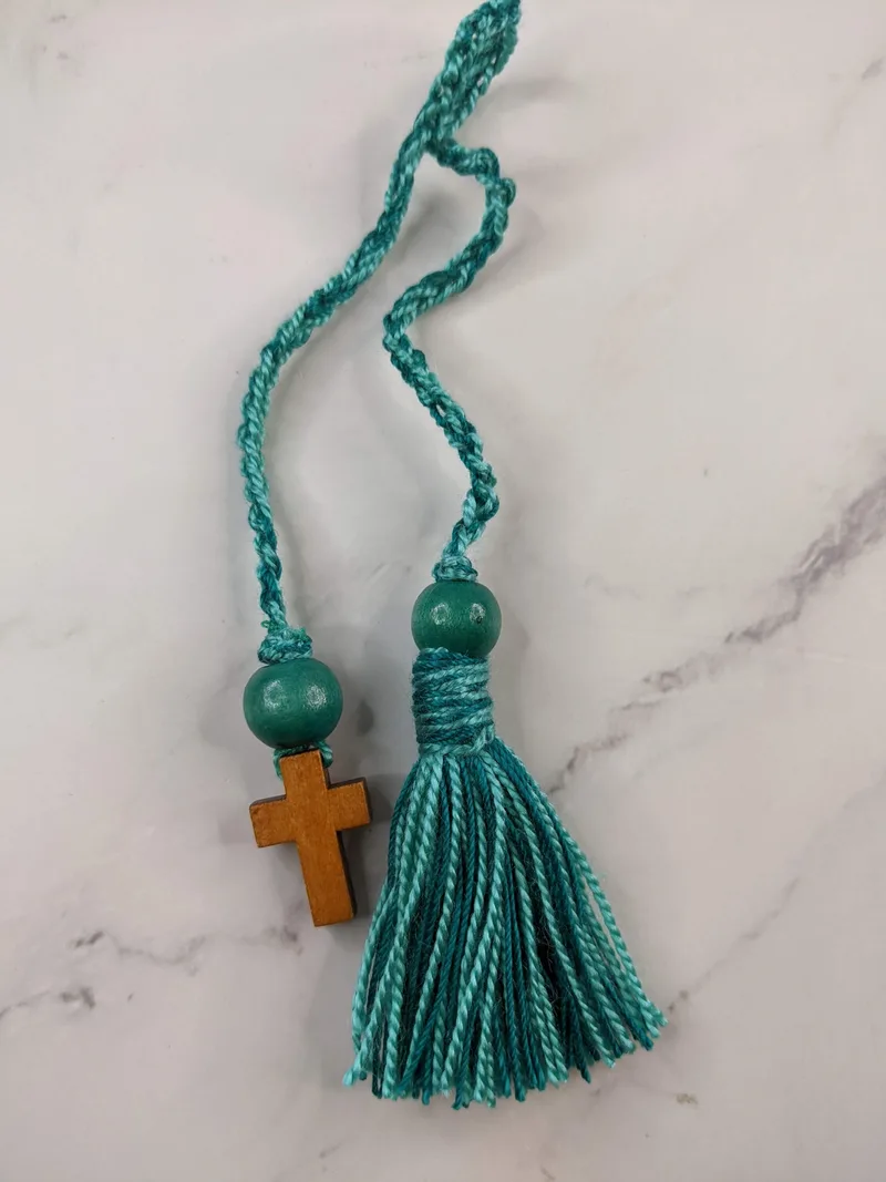 A crocheted teal bookmark with a wooden cross charm and a teal string tassel.