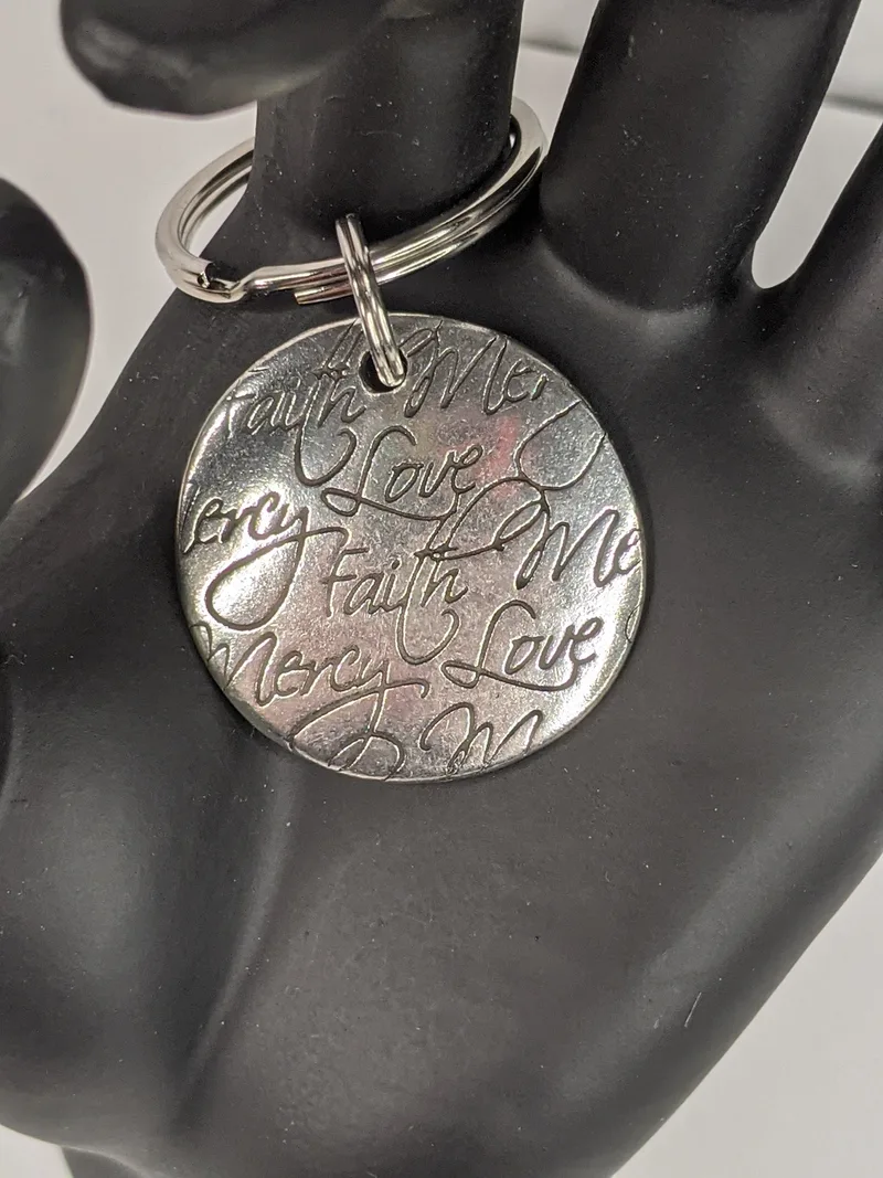 A silver metal keyring featuring the words Love, Faith and Mercy.