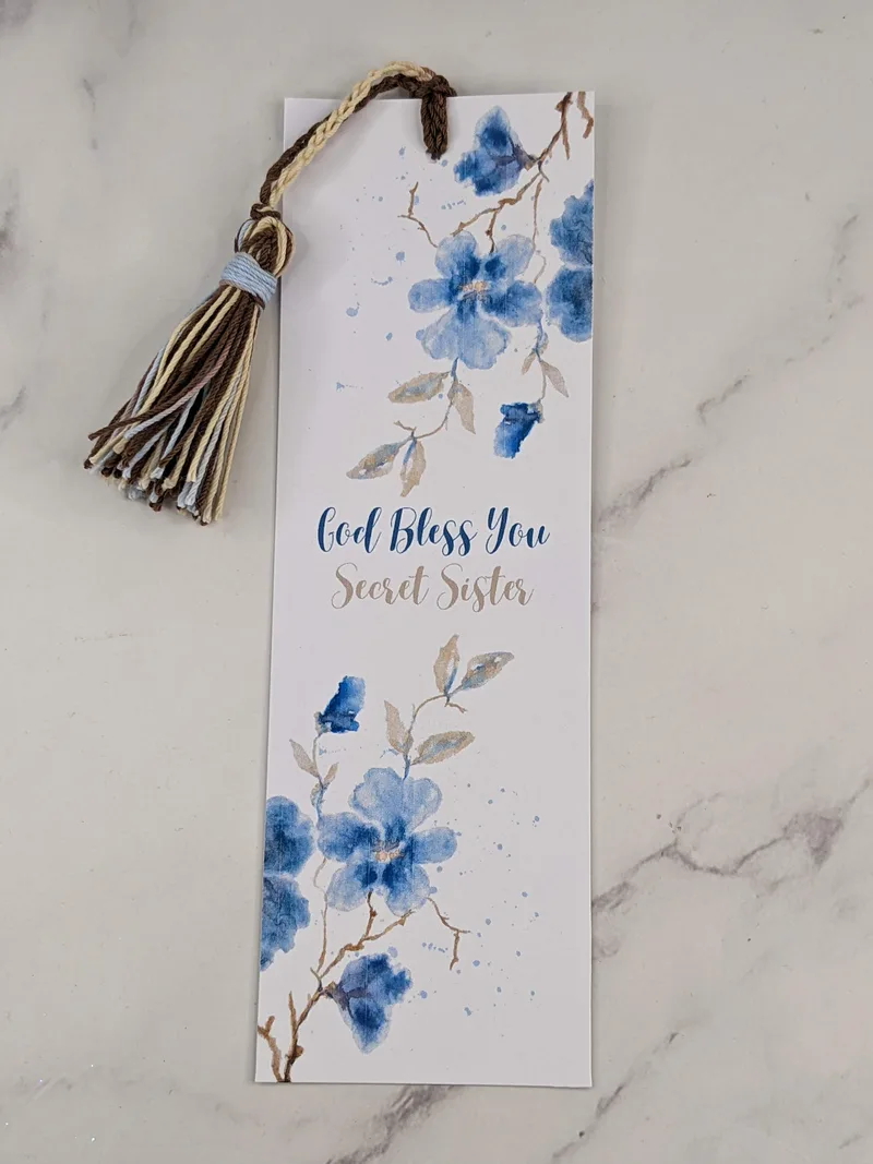 A Secret Sister Blessing bookmark with a white background, a design of blue flowers, and a matching string tassel.