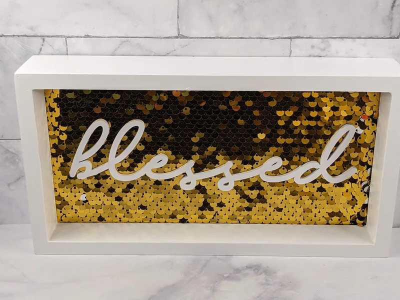 A golden sequin wall decor sign with the word "blessed" in white lettering, surrounded by a white frame.