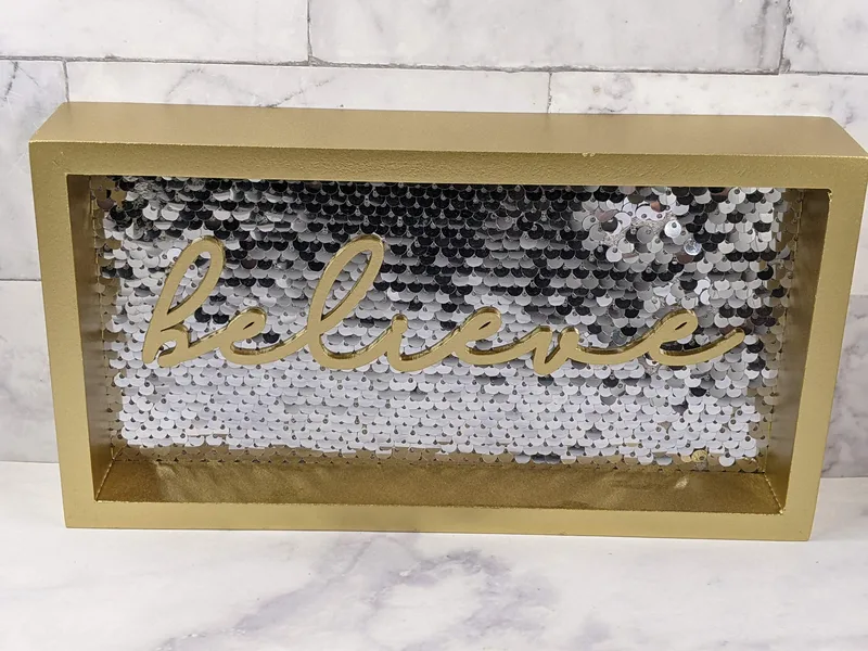 A wall decor sign with a background of silver sequins, featuring the word "believe" in gold lettering, surrounded by a gold frame.