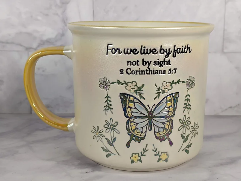 A religious gift mug with a blue butterfly design and a Bible verse about living by faith, not by sight.