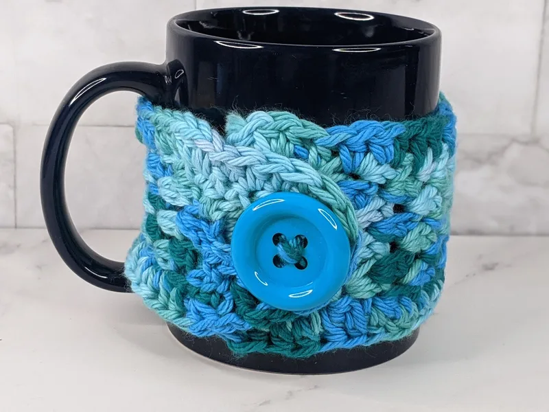 A handmade crocheted aqua blue mug cozy with a blue button closure.