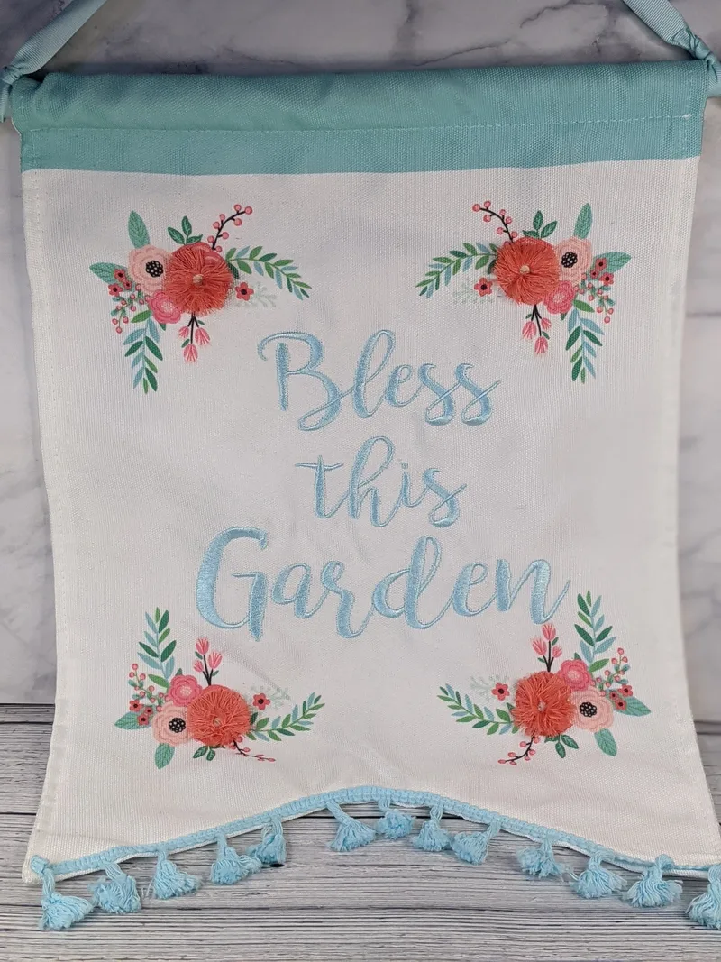 A wall banner that is white fabric with pink flowers embroidered and the words "Bless this garden" in blue.