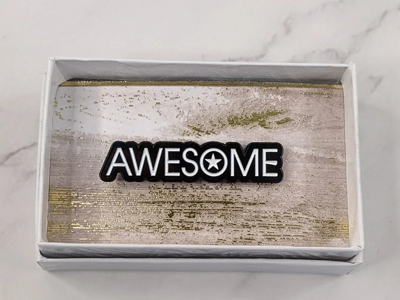 A black lapel pin with white lettering, featuring the word "awesome."