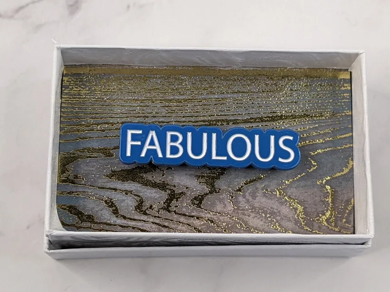 A blue lapel pin with white lettering, featuring the word "fabulous."