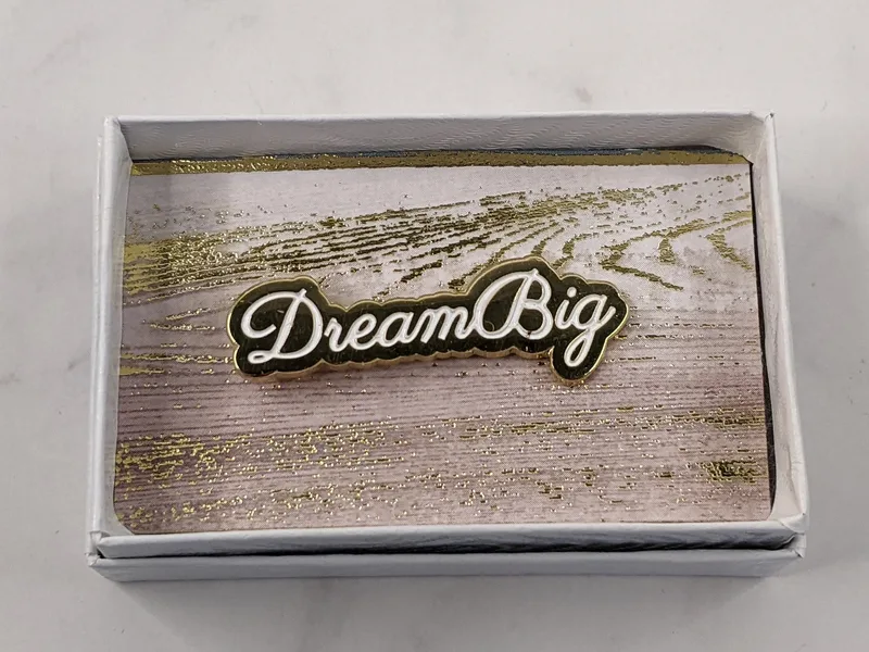 A gold lapel pin with white letters featuring the words, "Dream Big."