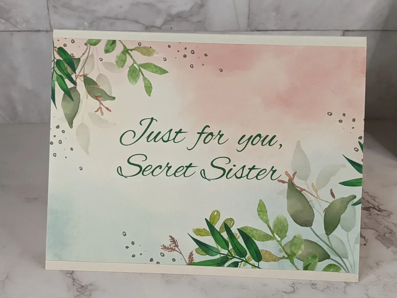 A greeting card for a Secret Sister, wishing her an amazing day, with a pink watercolor design and a green leaf pattern.