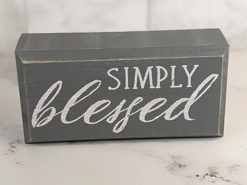 A gray wooden sign that sits on a shelf, with white painted lettering that says, "simply blessed."