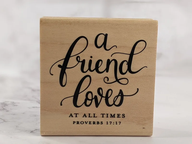 A religious rubber stamp mounted on a wooden block with the Bible verse: A friend loves at all times.