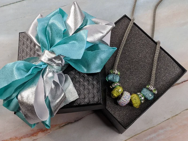A metal rope necklace with aqua blue and green slider beads, in a black jewelry box with a blue and silver bow.