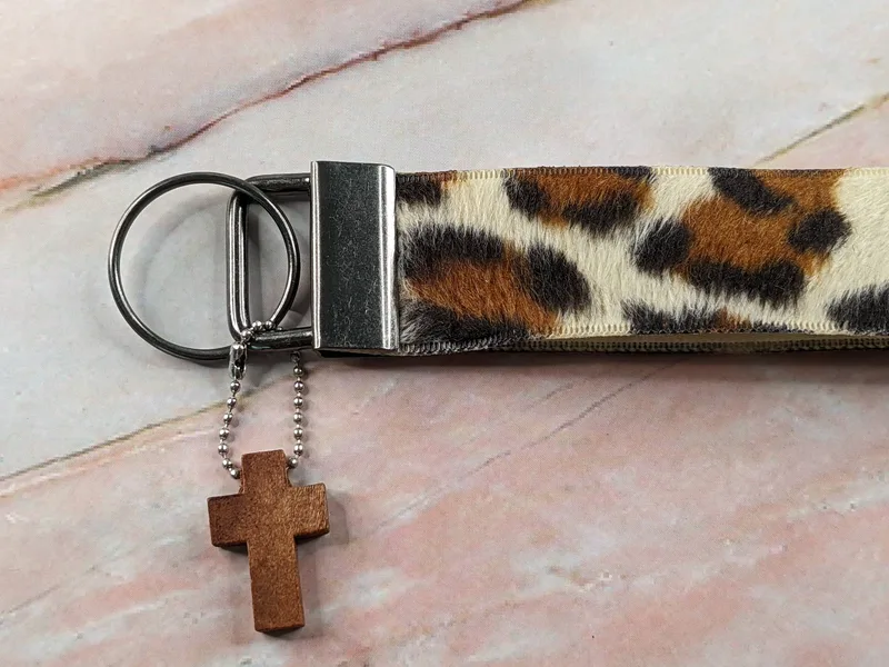 A wristlet keyring made of fuzzy fabric in shades of brown, tan and black, with a leopard-print and a wooden cross charm.