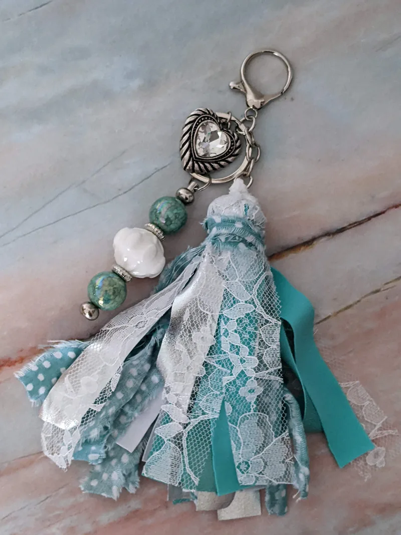 An aqua, silver, and white bag charm with a large fabric tassel and beaded charm stick with a heart pendant.