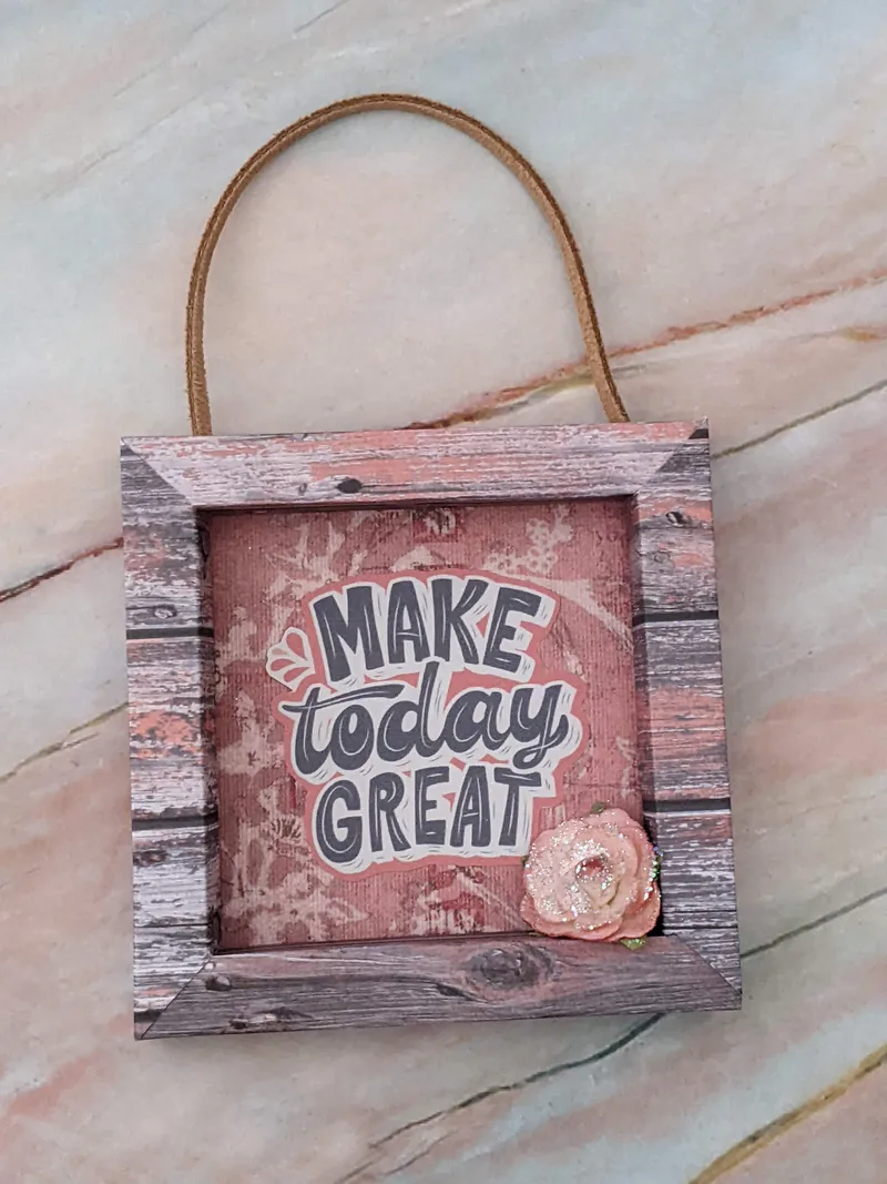 A paper wall sign with a framed look and a leather cord for hanging, with the words "make today great" and a glittery pink rose decoration.
