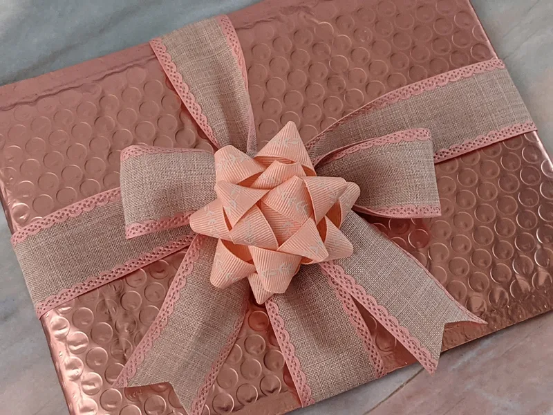 A coral metallic bubble mailer with a handmade bow topper, promoting a "Faith" surprise mystery bag.