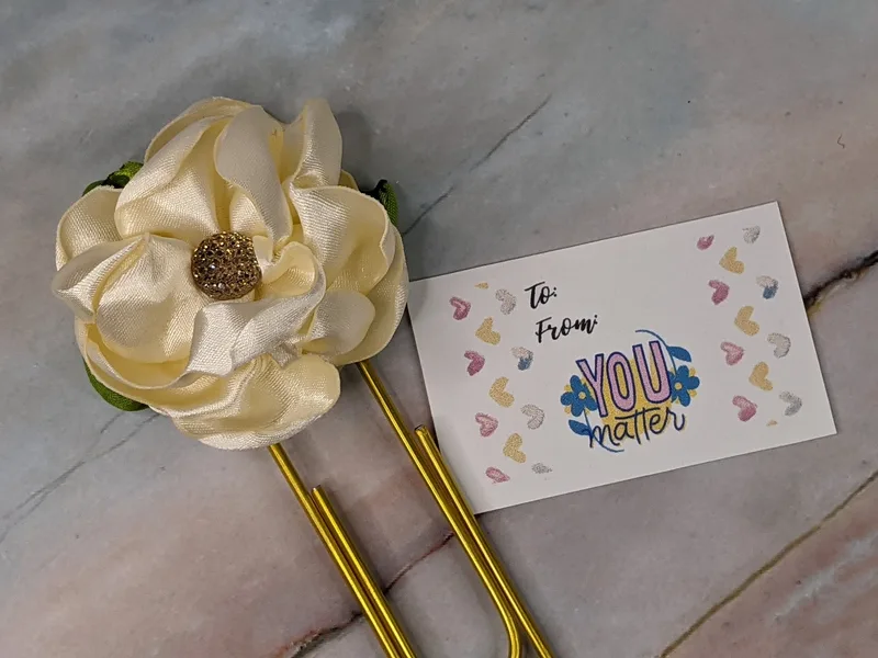 A cream-colored handmade satin flower on a gold paper clip with a tag that states "You matter."