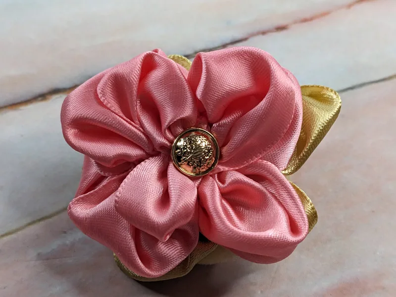 A handmade pink satin flower with a gold button center, mounted to a clip that attaches to a wrapped gift.