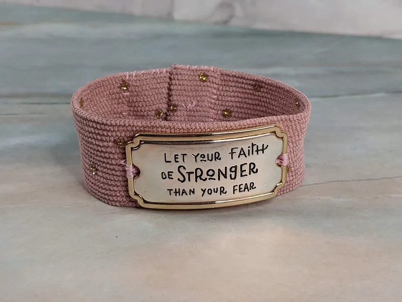 A pink corduroy bracelet with a "Let your faith be stronger than your fear" metal connector, golden glittery dots and an elastic-around-a-button closure.