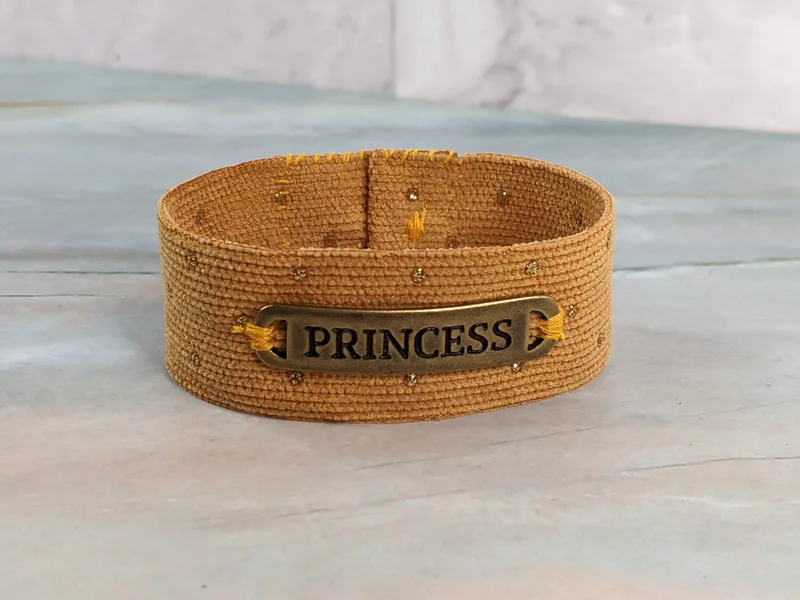 A gold corduroy fabric cuff bracelet with a gold button closure and a golden metal "princess" pendant tag.