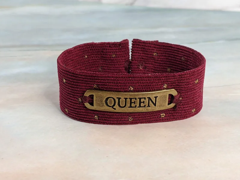 A burgundy corduroy cuff bracelet with a button clasp and a gold "queen" metal pendant tag.