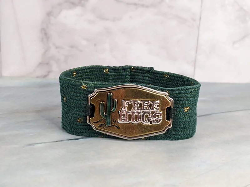 A green corduroy fabric cuff bracelet with a gold button closure and a "free hugs" gold metal pendant tag.