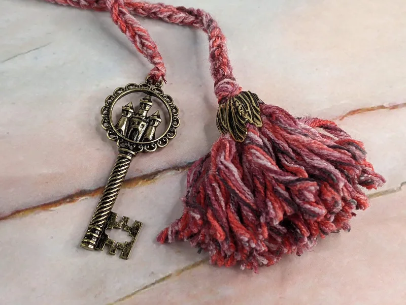 A dark pink yarn crocheted bookmark with a matching yarn tassel and a dark gold pendant shaped like a key with a castle symbol on it.