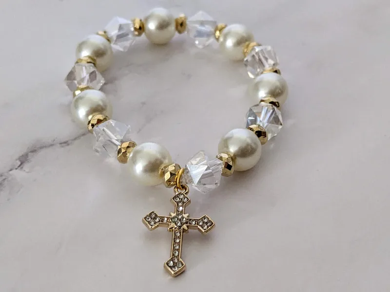 A handmade stretch bracelet made with pearl and glass beads, with a gold cross charm with rhinestones.